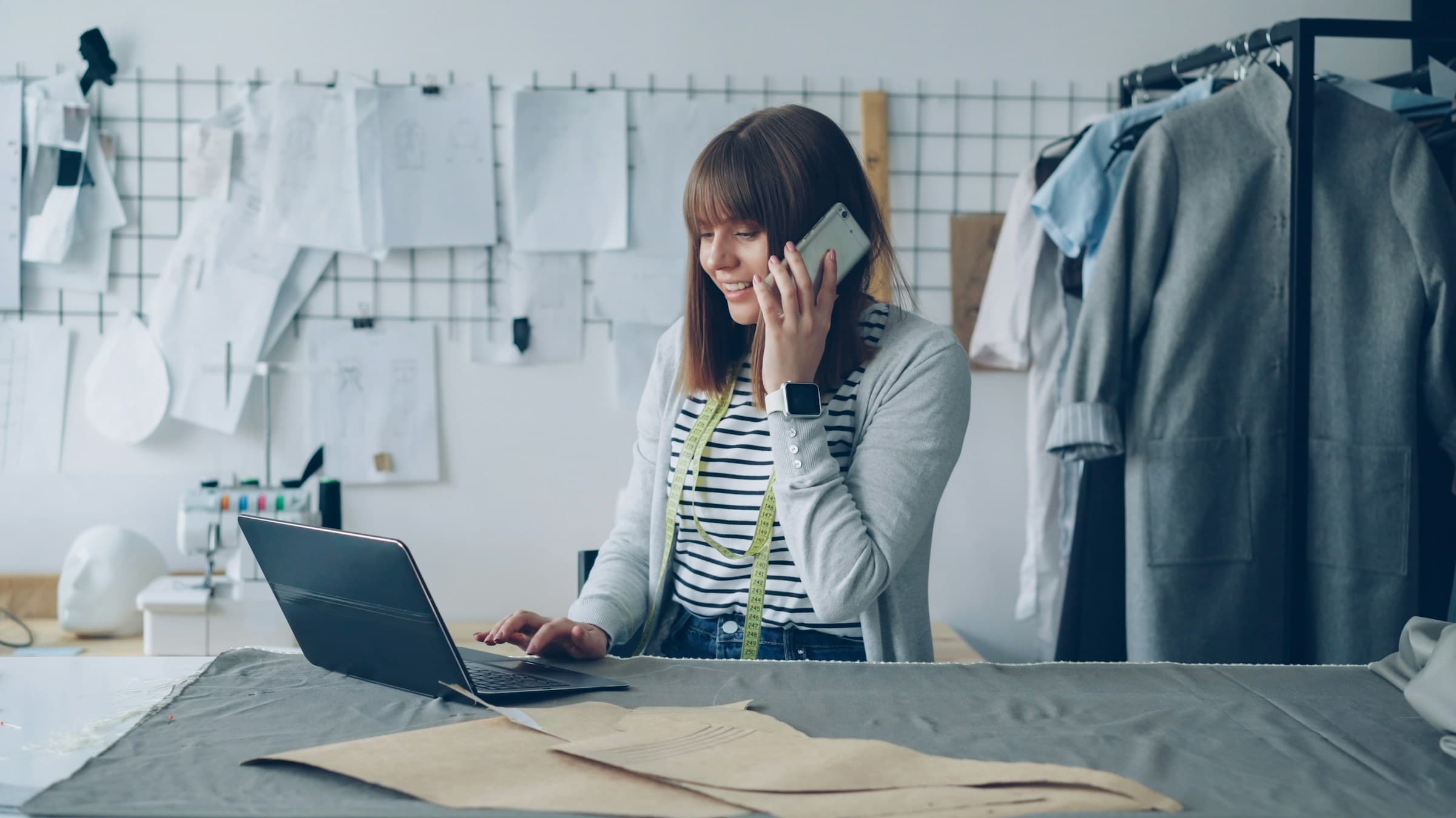 Fashion designer working on a laptop while talking on a phone, surrounded by their craft